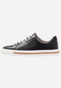 Baskets en cuir noir avec une silhouette basse, semelle en caoutchouc blanc, lacets blancs et accents beige sur le talon et la couture latérale.