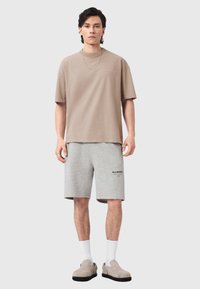 Μπεζ βαμβακερό t-shirt με κοντά μανίκια, συνδυασμένο με γκρι βαμβακερά σορτς με τυπωμένο λογότυπο. Το μοντέλο φοράει λευκές κάλτσες και γκρι παπούτσια τύπου slip-on.