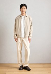Beżowa kurtka zapinana na zamek na białej koszulce, w połączeniu z kremowymi spodniami i ciemnobrązowymi butami na wsuwkę. Gładki materiał z subtelnymi teksturami.