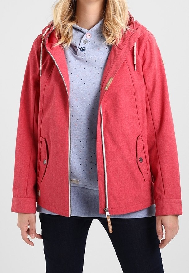 Femme portant une veste rouge à capuche sur une chemise bleu clair boutonnée avec de petits imprimés en forme de cœur, associée à un pantalon foncé.