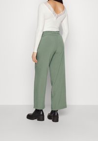 Gröna vida byxor med elastisk midja, i kombination med en vit långärmad topp med V-ringning och spetsdetaljer. Svarta klumpiga klackstövlar.