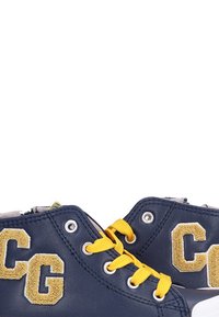 Zapatillas azul marino con cordones amarillos y parches "CG" en color beige en los laterales, mostrando un primer plano de los ojales y los detalles de costura.