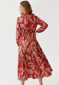 Abito rosso con maniche lunghe, vita stretta e gonna fluida. Presenta un motivo astratto di foglie bianche e un colletto.