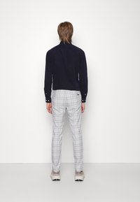 Man som står med ryggen mot kameran, iförd en marinblå långärmad skjorta, ljusgrå rutiga byxor och beige sneakers, stående mot en vit bakgrund.