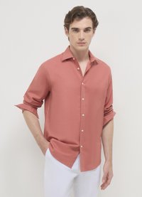 Chemise en corail clair boutonnée avec une coupe décontractée, manches longues retroussées aux poignets, et une texture lisse, associée à un pantalon blanc.