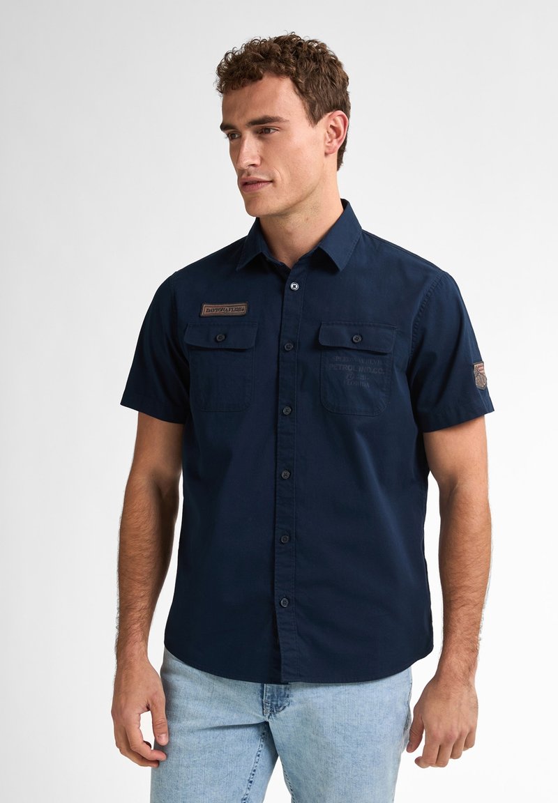 Camicia blu navy a maniche corte con due tasche frontali, colletto abbottonato e una toppa in tessuto sul petto. Texture liscia, design casual.