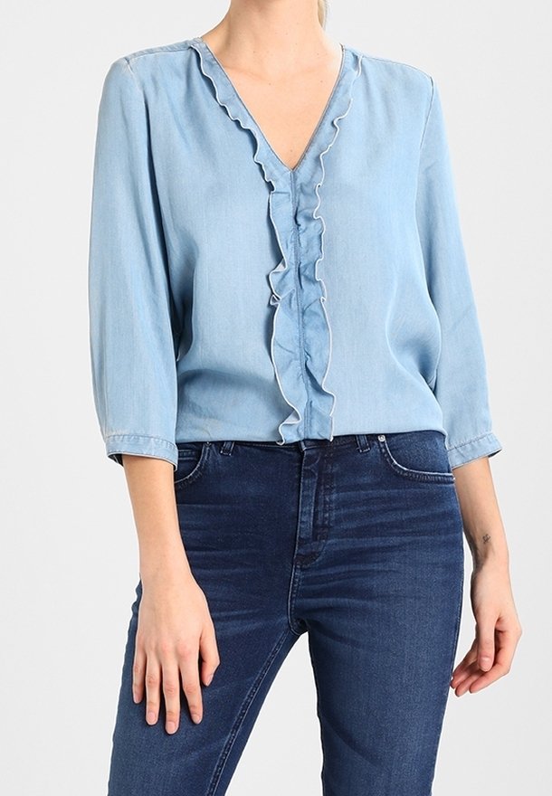 Helle blaue, denimartige Bluse mit V-Ausschnitt und Rüsche am Rand; drei Viertelärmel; glatte Textur; kombiniert mit dunkelblauen Jeans.