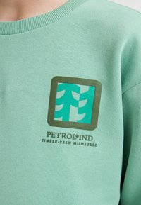 Sweatshirt vert clair à col rond avec un logo rectangulaire comportant des arbres stylisés en bleu canard et un nom de marque en marron en dessous.
