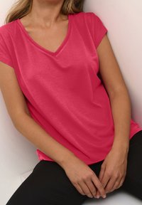 Top a maniche corte, con scollo a V, di colore rosa brillante, realizzato in tessuto liscio. Presenta una vestibilità rilassata con cuciture laterali. Senza fantasie né hardware visibile.