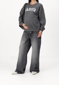 Sudadera gris con el estampado "DAKOTA", combinada con pantalones anchos gris. Tacones blancos puntiagudos. La modelo se sostiene el vientre, mostrando un atuendo casual.