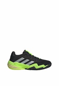 Zapatilla deportiva negra con franjas plateadas, acentos en verde neón y parte superior de malla texturizada, con suela de goma para mayor tracción.