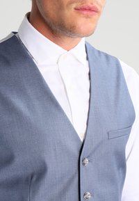 Chaleco gris texturizado con un diseño de escote en V, que presenta dos botones plateados y un bolsillo, llevado sobre una camisa blanca con cuello.
