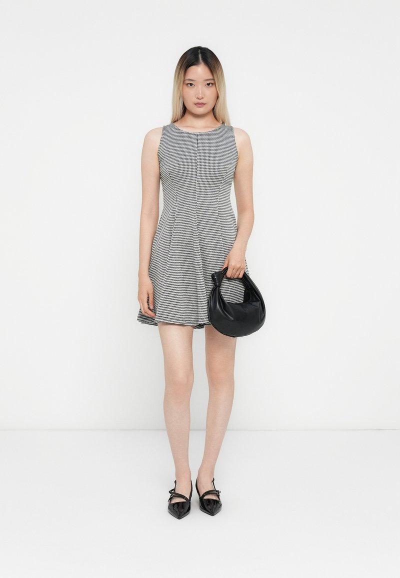 Robe sans manches gris et blanc à carreaux avec une jupe évasée. Associée à un sac à main circulaire noir et des ballerines noires à bout pointu.