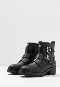 Sorte lær ankle boots med to sølvspenner, glidelåser på siden, en kraftig såle og en moderat blokkhel. Teksturet overdel med stikningsdetaljer.