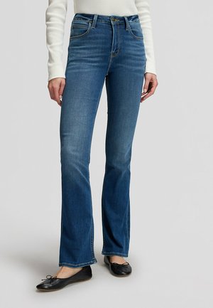 Person wearing blue bootcut jeans and black ballet flats, standing against a plain light background.