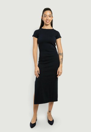 Giovane donna in piedi, indossa un abito midi nero aderente a maniche corte e ballerine nere, con tatuaggi sul braccio sinistro e sulla caviglia destra.