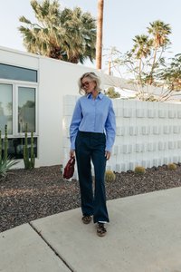 Košilové tričko s modrými vertikálními pruhy, tmavé široké kalhoty, boty s leopardím potiskem a červená kabelka. Texturovaná bílá zeď v pozadí.