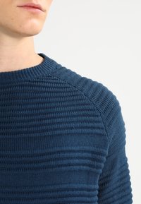 Dunkelblauer Strickpullover mit gerippter Textur, rundem Ausschnitt und markanten horizontalen Streifen über dem Stoff.
