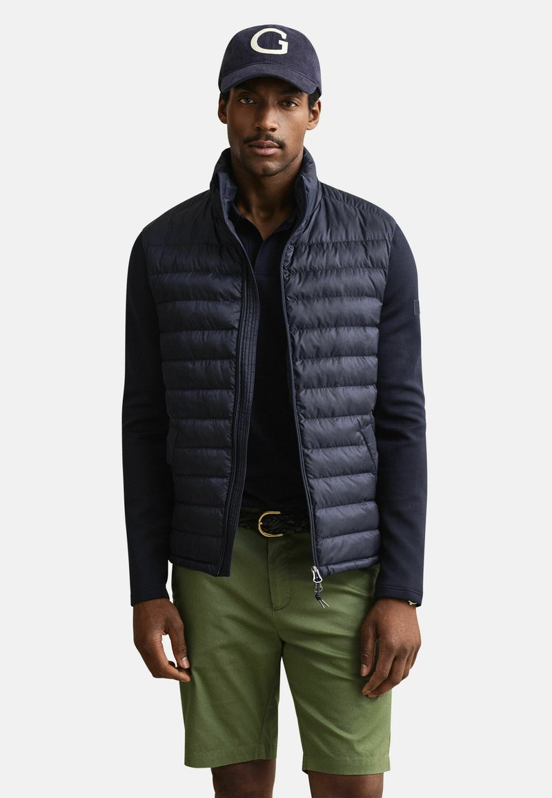 Mann trägt marineblaue Mütze mit „G“, marineblaue gesteppte Weste über langärmeligem Hemd und olivgrüne Shorts, steht vor schlichten Hintergrund.