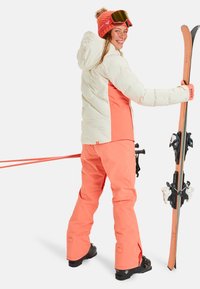 Skiausrüstung mit einer weißen und korallenfarbenen Kammmanteljacke, korallenfarbener Hose und schwarzen Skistiefeln. Inklusive orangefarbener Mütze und Skibrille, hält Skier.