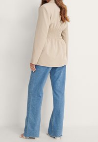 Beżowa marynarka o dopasowanym kroju z zaszewkami z tyłu i gładką fakturą, zestawiona z jasnoniebieskimi spodniami typu wide-leg z postrzępionymi brzegami.