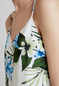 Blommig ärmlös topp med gröna och blå blommor på en vit bakgrund. Materialet verkar lätt, med tunna axelband.