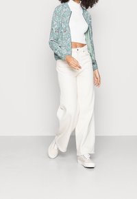 Vrouw in een witte cropped coltrui, lichtblauwe bloemen bomberjack, wijde crèmekleurige broek en witte sneakers die over een lichte vloer loopt.