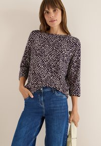 Långärmad, mörklila topp med ett krämfärgat abstrakt mönster, tillsammans med blå denimjeans. Tyget verkar mjukt och texturerat.