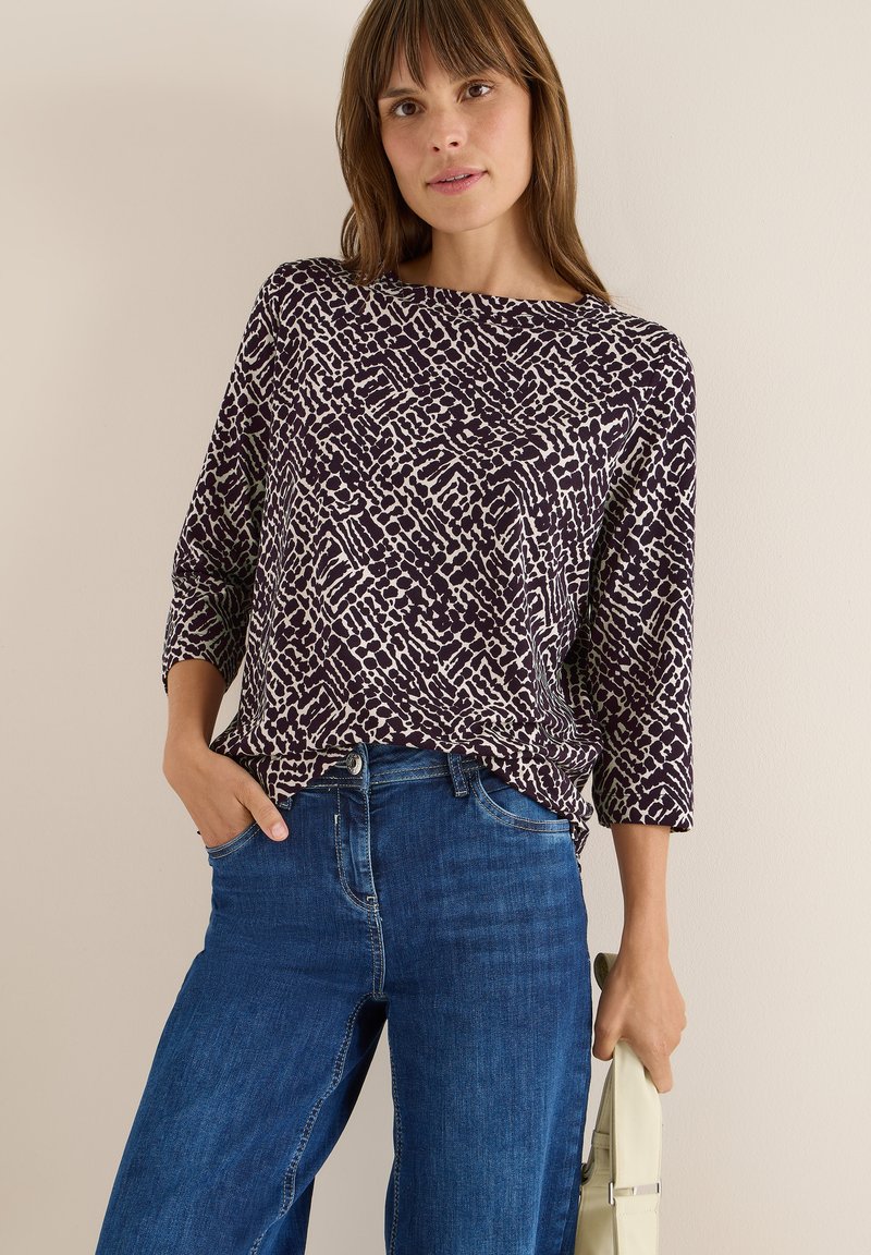 Långärmad, mörklila topp med ett krämfärgat abstrakt mönster, tillsammans med blå denimjeans. Tyget verkar mjukt och texturerat.