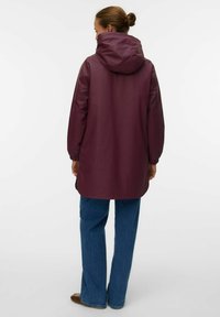 Giacca impermeabile bordeaux con cappuccio, maniche lunghe e polsini elasticizzati; indossata sopra jeans a zampa d'elefante blu, che mostrano una texture liscia e raffinata.