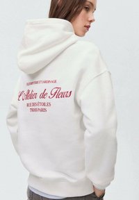 Hoodie bianco oversize realizzato in morbido tessuto. Presenta un testo ricamato rosso sul retro, comprensivo del nome del marchio e dei dettagli dell'indirizzo.