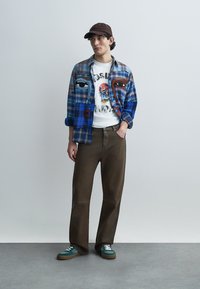 Chemise à carreaux tons bleu et marron portée sur un t-shirt blanc à motif ; pantalon marron à jambes larges ; baskets vertes avec lacets blancs ; casquette marron.