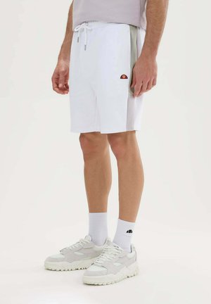 Hombre con pantalones cortos blancos con cordón y logo, calcetines blancos con logo y zapatillas blancas, de pie sobre un fondo liso y claro.