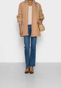 Veste matelassée beige avec un col chemise, associée à un haut en tricot crème, un jean flare bleu et des bottines marron. Petit sac beige.