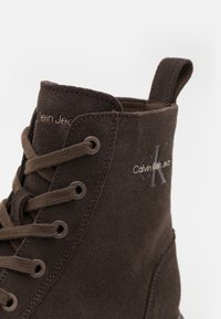 Stivale alla caviglia in suede marrone con design a lacci, dotato di un inserto a occhio e branding discreto sulla parte superiore. Finitura liscia e testurizzata.