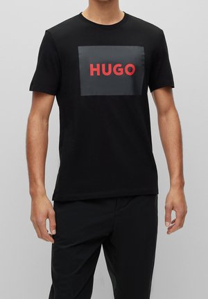 Mand iført en sort T-shirt med korte ærmer med en grå firkant og rød "HUGO" tekst på brystet, kombineret med sorte bukser.
