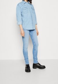 Camisa de denim azul claro con dos bolsillos en el pecho y mangas enrolladas, combinada con jeans ajustados azul claro y botas chunky negras con cordones.