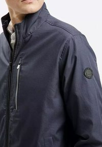 Granatowa kurtka z wysokim kołnierzem, zamek błyskawiczny z przodu i kieszeń na zamek błyskawiczny na piersi. Okrąły logo na rękawie dodaje detalu.