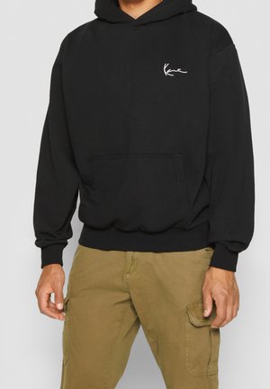 Man met zwarte hoodie met klein wit logo op de borst en beige cargobroek, staand tegen een neutrale achtergrond.