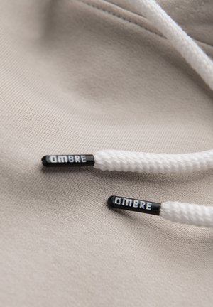 Cordon de sweat à capuche blanc avec embouts noirs étiquetés "OMBRE" reposant sur un fond en tissu beige.
