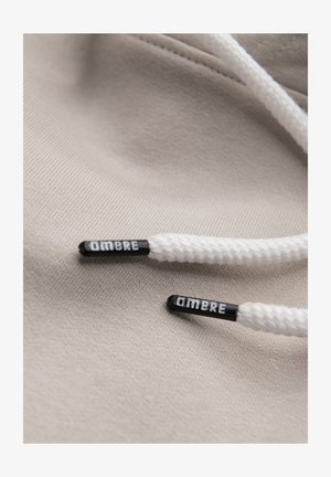 Cordon de sweat à capuche blanc avec embouts noirs étiquetés "OMBRE" reposant sur un fond en tissu beige.