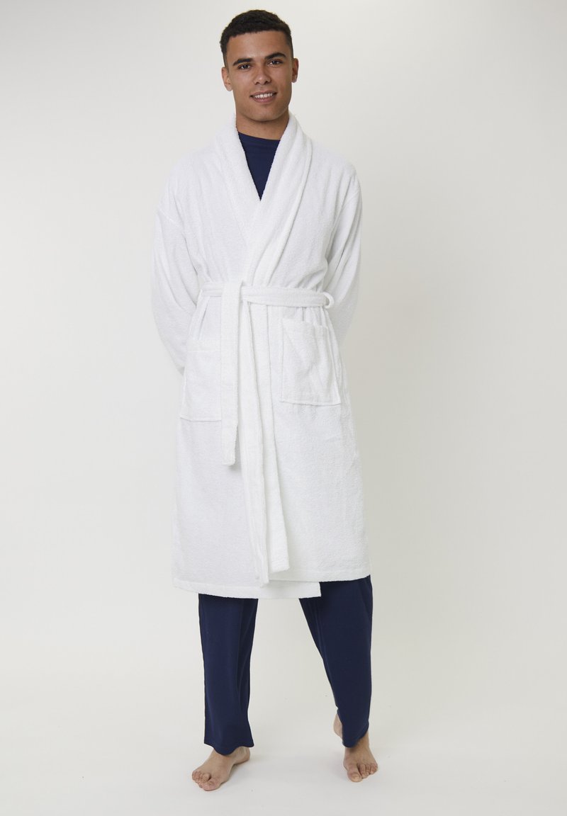 Un jeune homme souriant, portant un peignoir blanc par-dessus un pyjama bleu marine, se tenant pieds nus devant un fond clair.