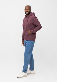 Bordeauxfarbener Kapuzenpullover mit einer Fronttasche und Kordelzug, kombiniert mit hellblauen Jeans und weißen Turnschuhen. Glatter Stoff, lässige Passform.