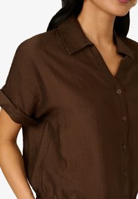 Chemise boutonnée marron avec un tissu texturé, des manches courtes et un col, ornée de lignes verticales subtiles et de boutons assortis.
