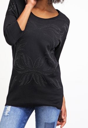 Frau trägt ein schwarzes Langarmshirt mit silbernem Nietenblumenmuster und blaue, ausgewaschene Jeans, Hand nahe am Gesicht, vor weißem Hintergrund.