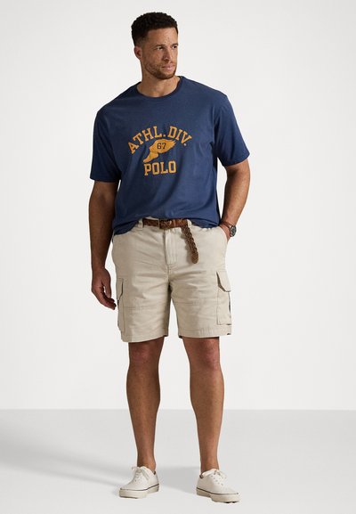 Tricou bleumarin cu imprimeu "ATHL.DIV. 67 POLO" în galben, asortat cu pantaloni scurți cargo bej, cu buzunare laterale și o curea împletită maro.