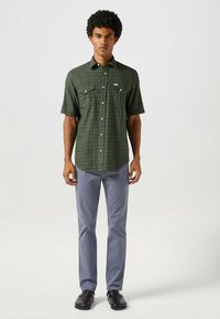 Chemise à manches courtes à carreaux verts avec deux poches poitrine, associée à un pantalon gris clair et des chaussures noires, se tenant sur un fond neutre.