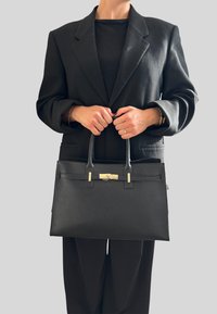 Czarna skórzana torebka o strukturalnym kształcie, teksturowanej powierzchni, złotych akcentach metalowych i dwóch uchwytach. Nosi się ją z czarnym żakietem.