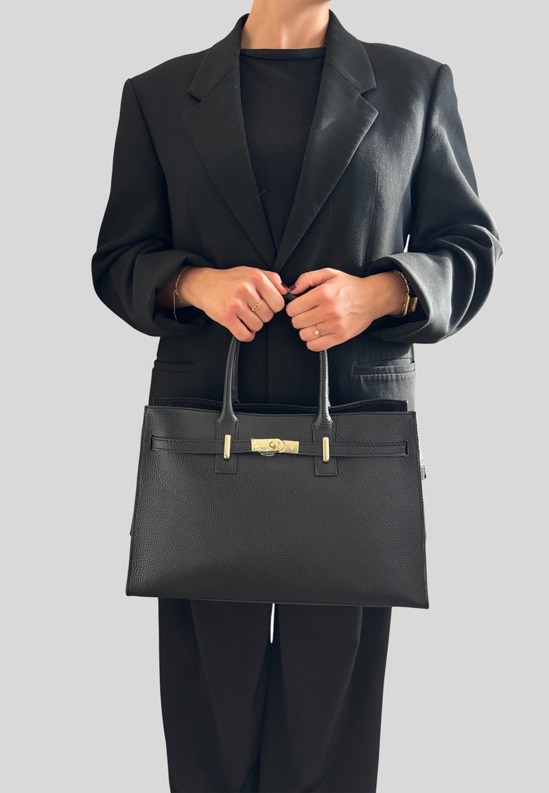 Czarna skórzana torebka o strukturalnym kształcie, teksturowanej powierzchni, złotych akcentach metalowych i dwóch uchwytach. Nosi się ją z czarnym żakietem.