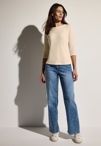 Beige strukturierter Langarm-Top, kombiniert mit hellblauen weit geschnittenen Jeans und weißen Sneakers. Einfaches Design, keine sichtbaren Muster.